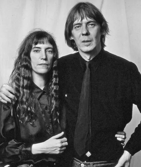 Seiji Matsumoto Black and white photo of Patti Smith with long dark hair, short fringe, in dark satin blouse, with her arm around her husband Fred's waist and his arm around her shoulder. Fred is wearing dark shirt and dark tie.