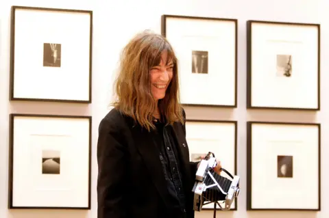 Getty Images Patti in black jacket and denim shirt, holding a camera and smiling in front of six prints of her artwork on the wall.