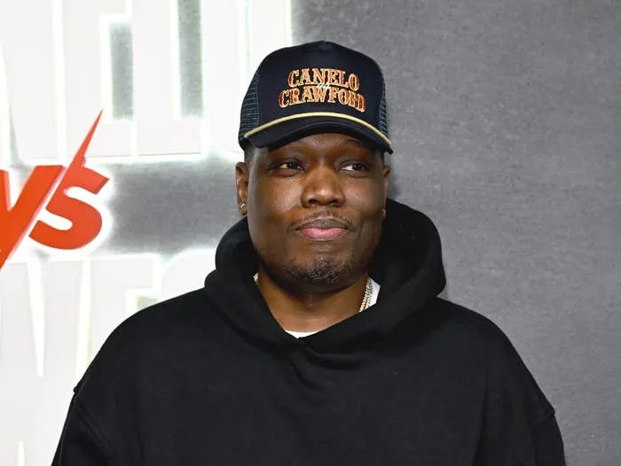 A man wearing a hoodie and a branded baseball cap stands on a red carpet with a neutral expression