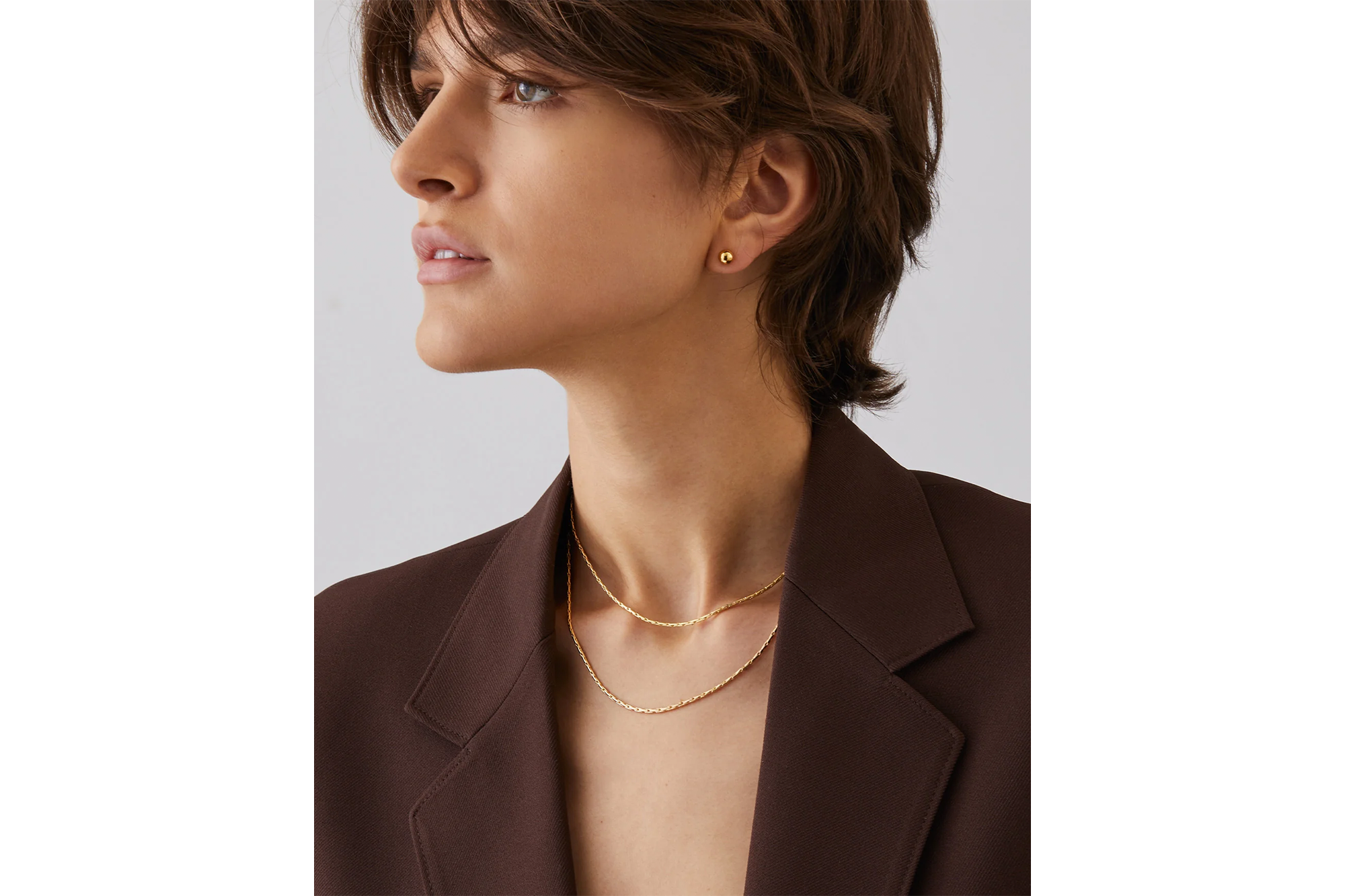 A woman in a brown blazer wearing a short gold chain necklace and a gold stud earring.