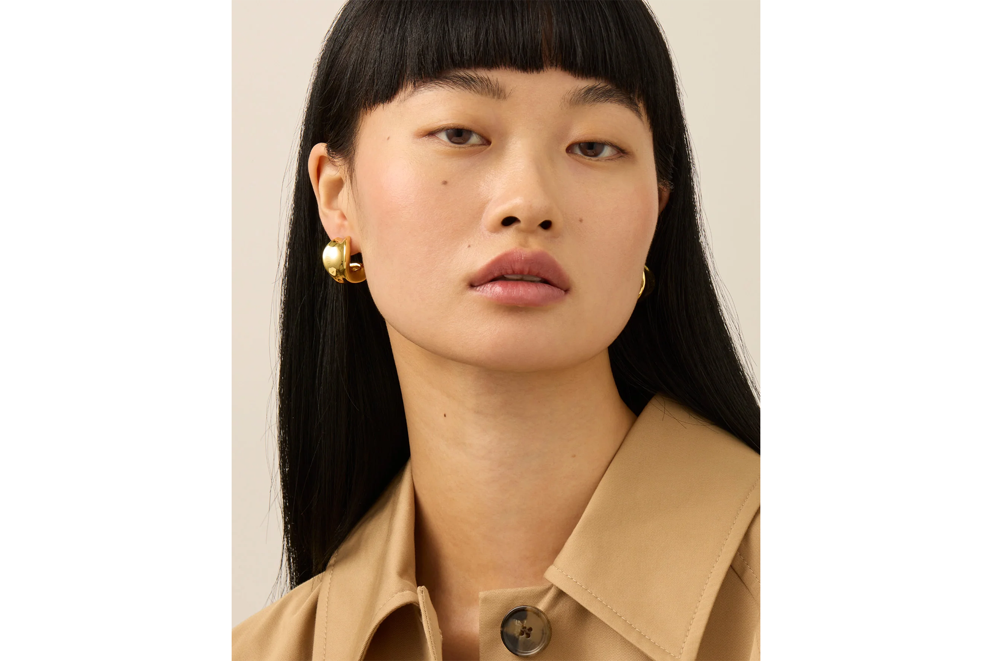 A person wearing large, shiny gold hoop earrings and a tan jacket.