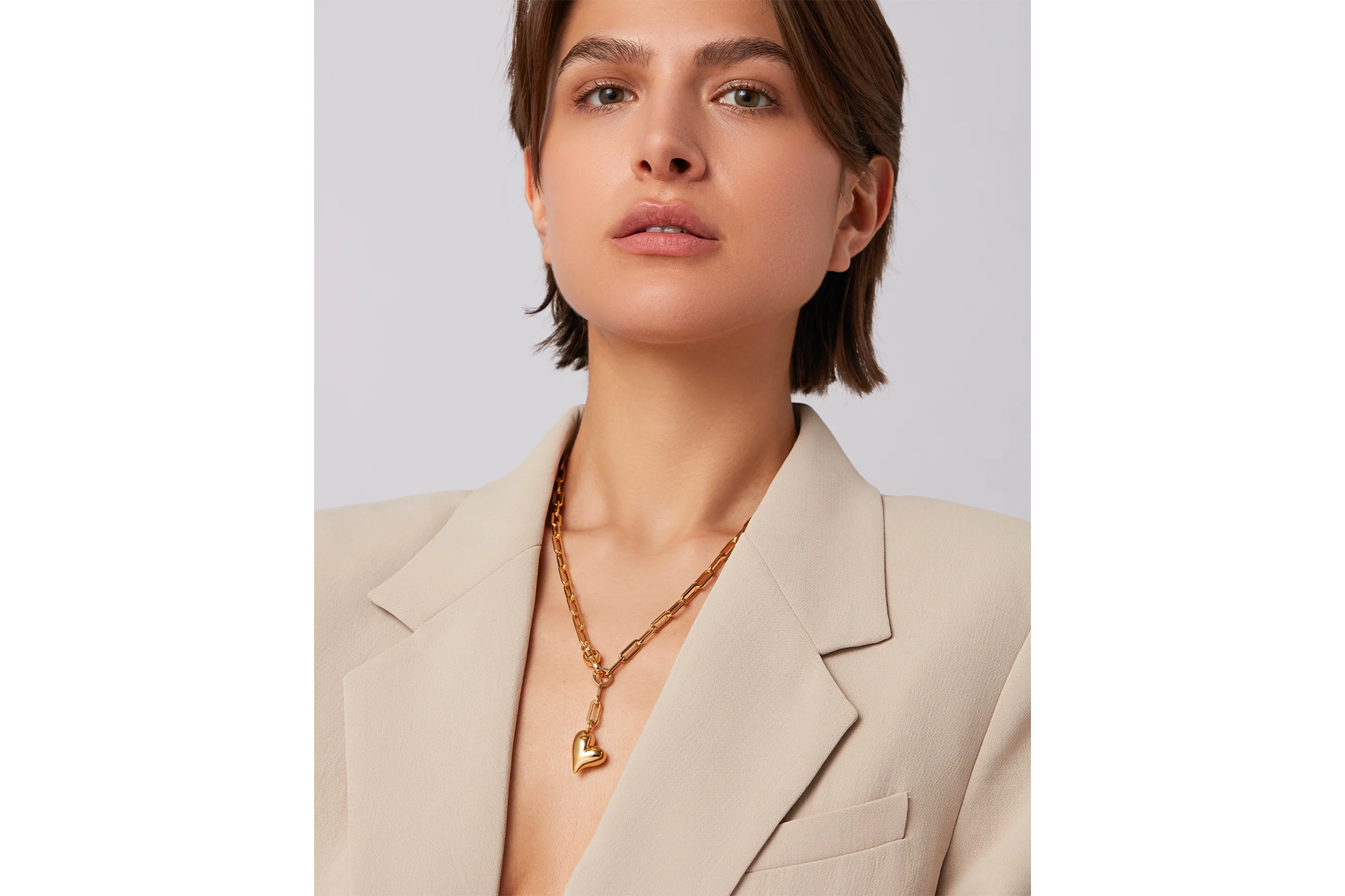 Woman with short brown hair wearing a beige blazer and a gold chain necklace with a heart pendant.