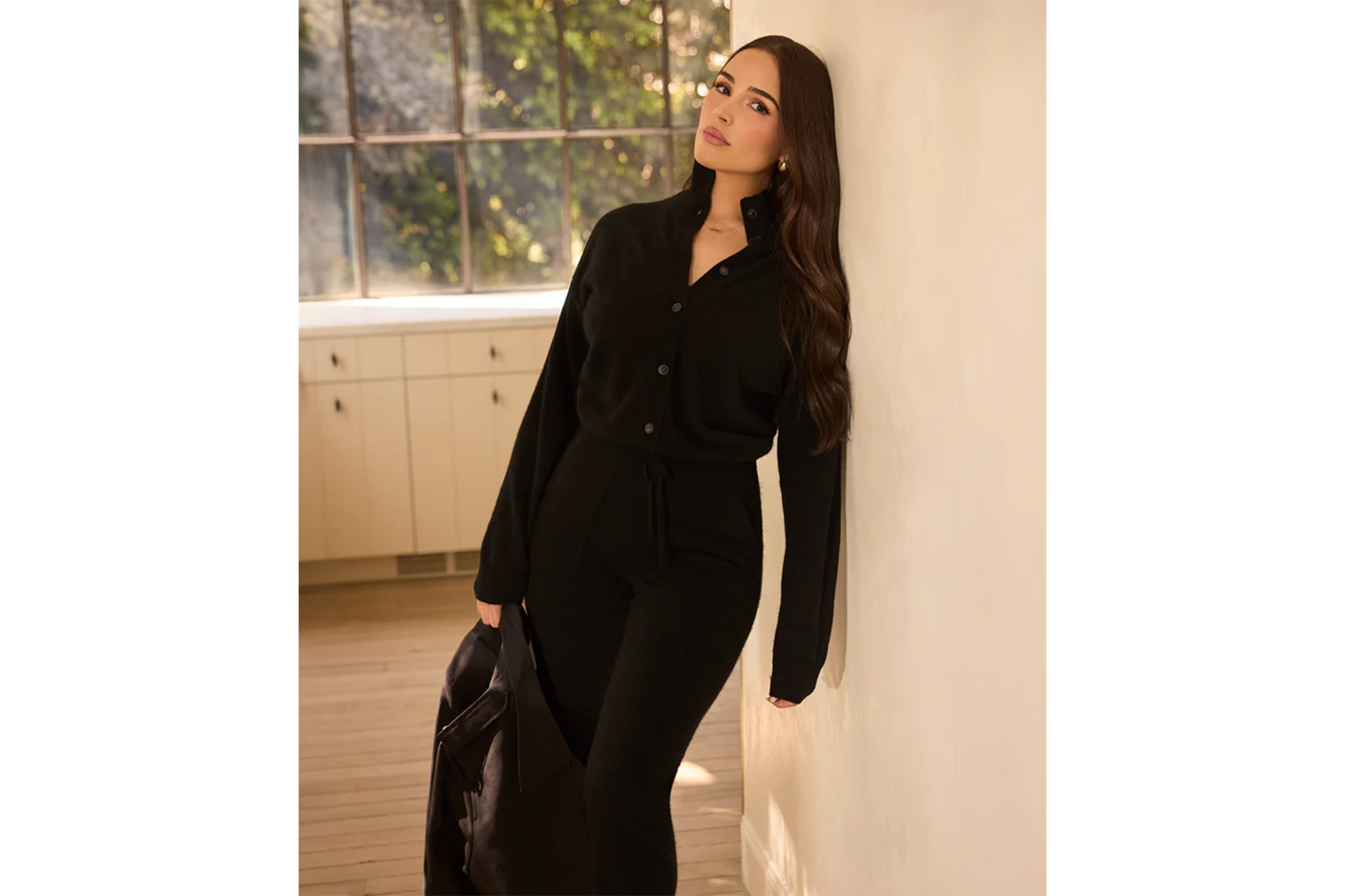 A woman in a black cardigan and matching pants holding a black jacket, leaning against a wall.