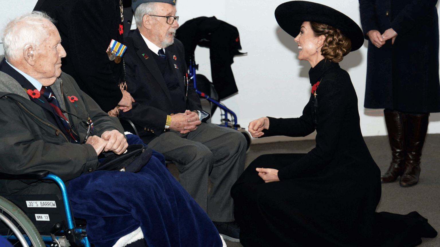 Princess Kate Admits Prince George “Knows More About” One Subject Than She Does During Sweet Chat With Veterans Kate Middleton kneeling in a black coat and hat talking to veterans in wheelchairs