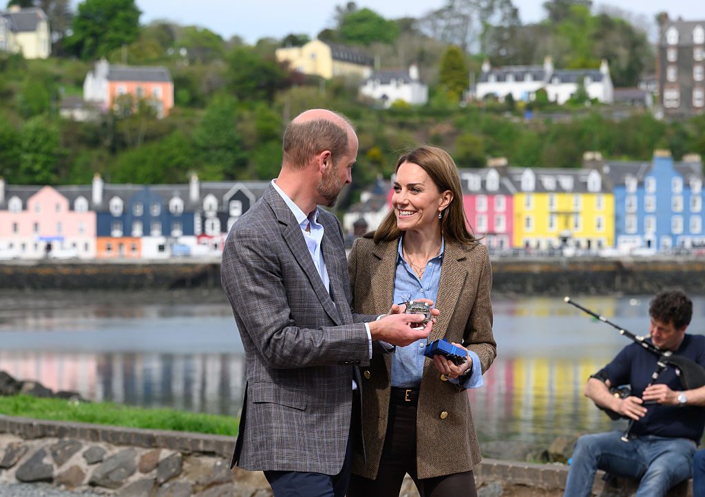 Prince and Princess of Wales with colourful houses and a bagpiper behind them in Tobermory