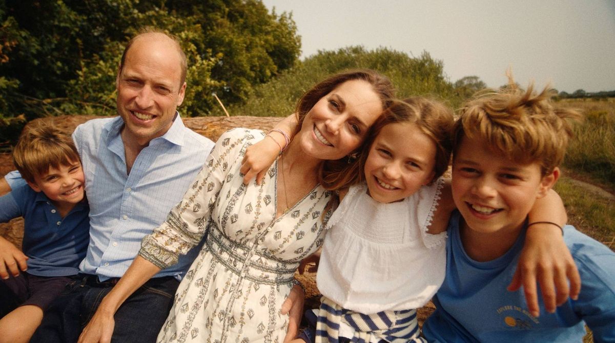 A family is gathered together for a photo, smiling and posing in a natural outdoor setting. A man wearing a light blue shirt sits beside a woman in a white dress with a pattern. To their left, a young boy in a dark shirt is partially visible. Next to the woman, a young girl in a white shirt stands next to a boy in a blue shirt, who is also smiling and looking at the camera. Trees and greenery are visible in the background.