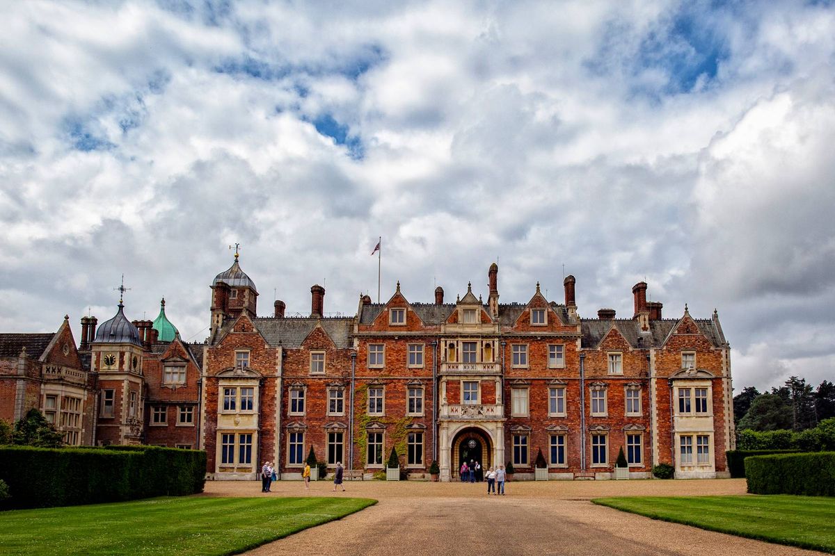 "A grand, historical building with numerous windows and turrets, set against a backdrop of a partly cloudy sky. The entrance is flanked by neatly trimmed hedges, leading to a wide driveway that extends into the distance."