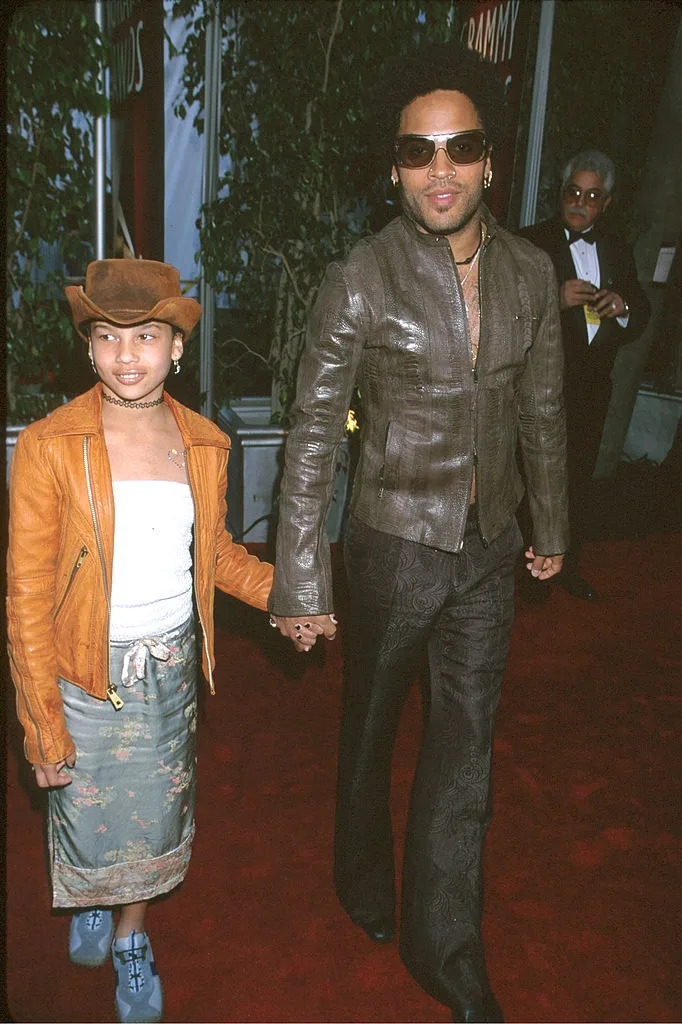 Two people on a red carpet; one in a leather jacket and wide pants, the other in a jacket and skirt, both holding hands and wearing hats