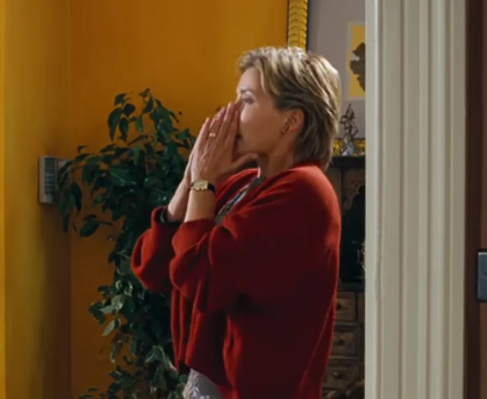 Person stands indoors, clasping hands over mouth in apparent surprise or concern, next to a potted plant