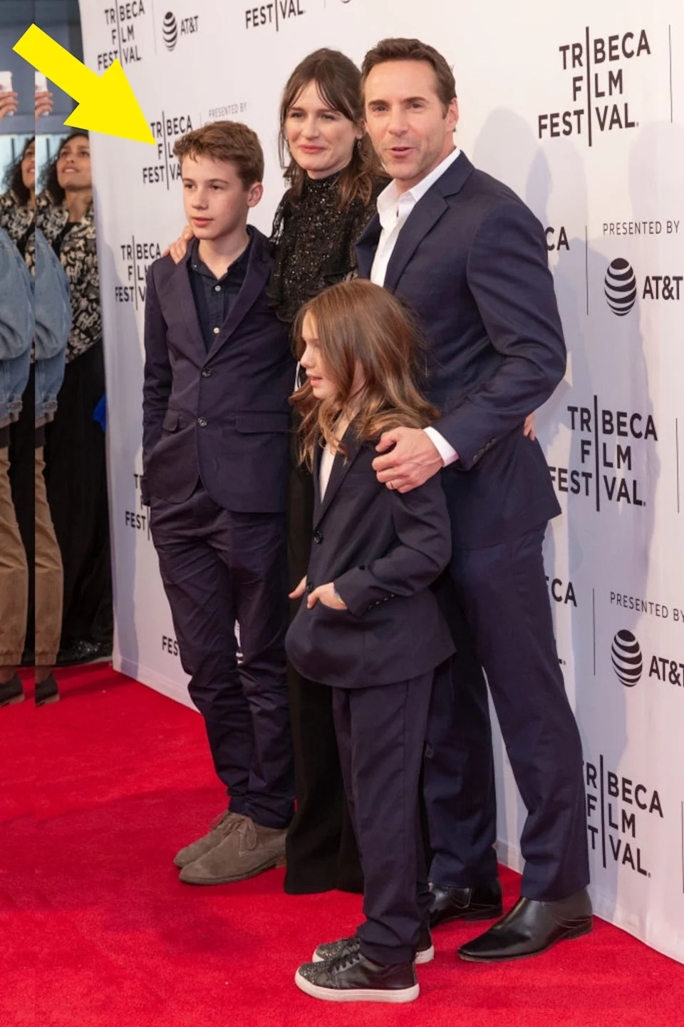 A group poses on the red carpet at the Tribeca Film Festival. The adults wear stylish formal wear, and the children are in suits with casual footwear