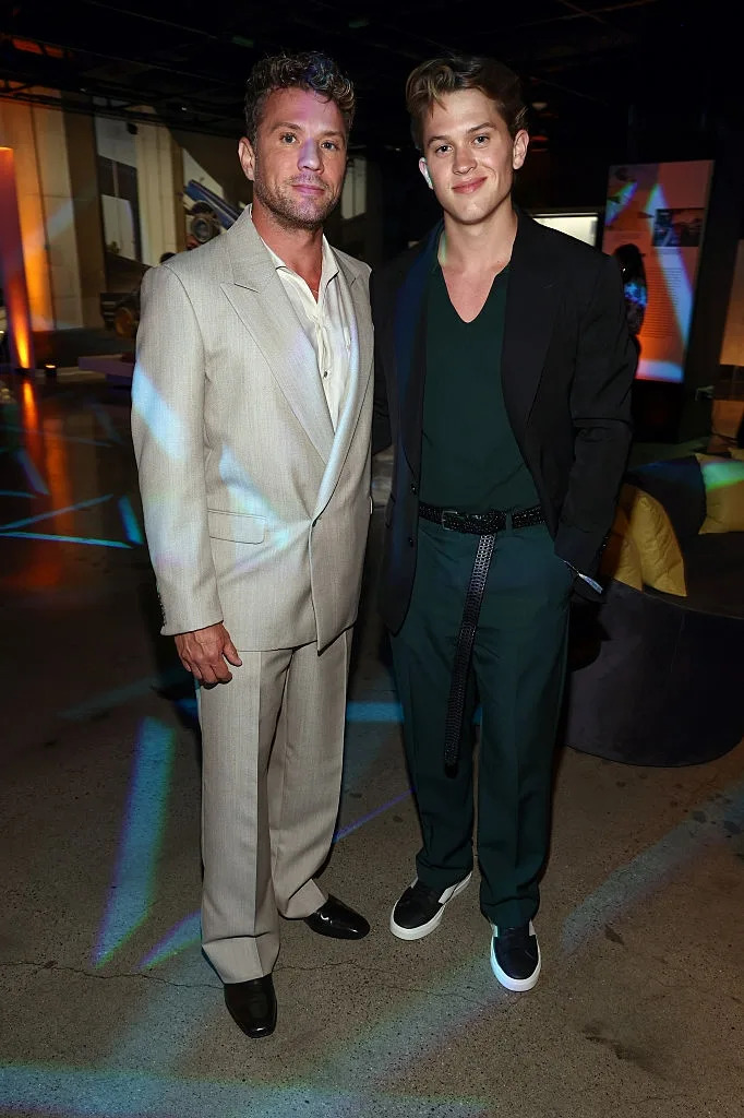 Two individuals on an event floor; one in a light suit with no tie, the other in a casual blazer with a green shirt and sneakers