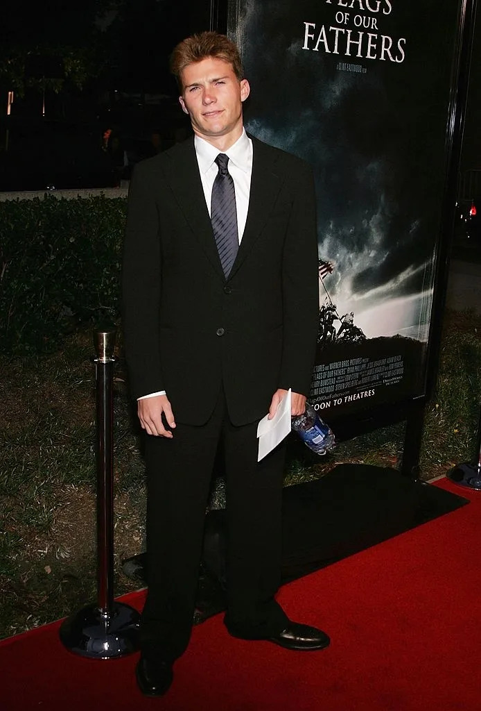 Person in a black suit and tie stands on a red carpet, holding a paper and water bottle, beside a movie poster