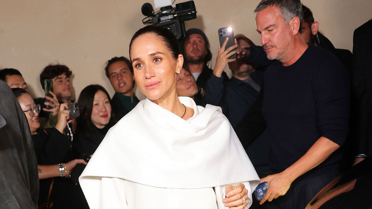 A close-up of Meghan Markle in white walking in front of photographers.