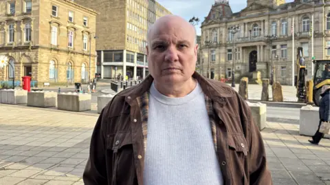 Gemma Dillon/BBC A bald man wearing a grey top and brown jacket is facing the camera. 
Behind him is Dewsbury Town Hall a stone building, and a boarded up building. 