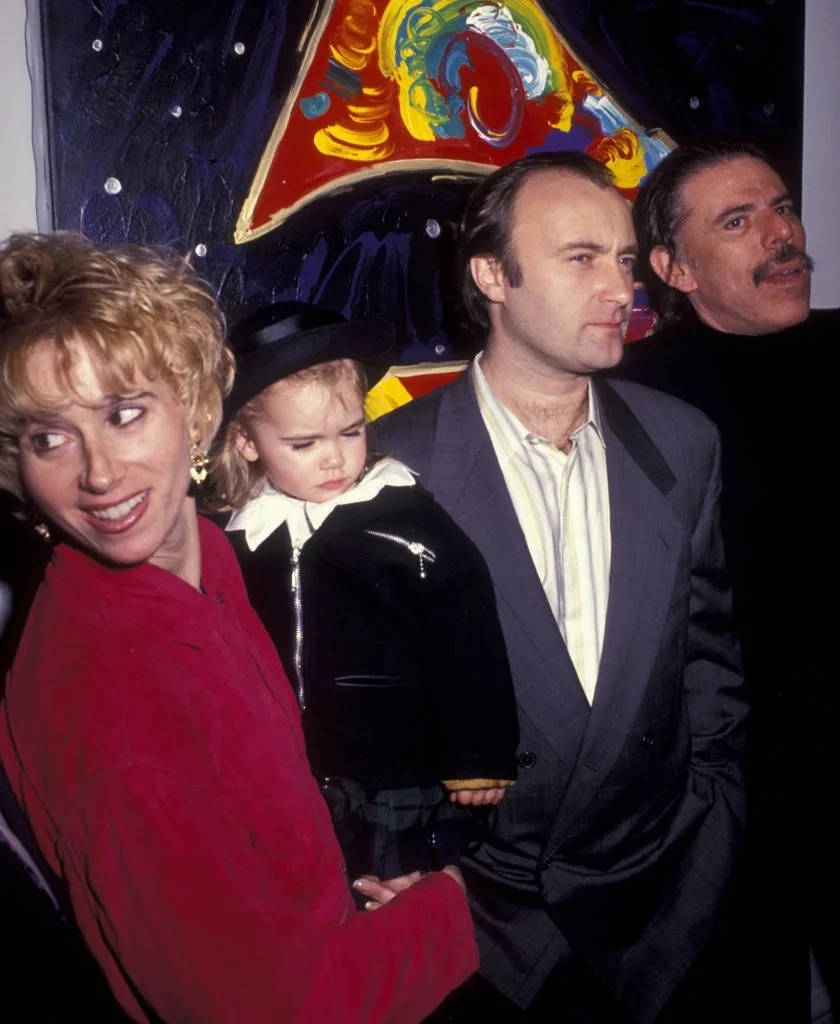 Group of four people standing together indoors; one adult holds a child wearing a hat. Artwork is visible in the background