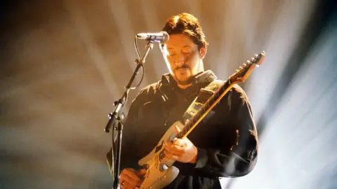 Getty Images Chris Rea performing on stage in 1990, playing an electric guitar and standing in front of a microphone. He is wearing a dark long-sleeved outfit, and bright stage lights create dramatic beams radiating in the background.