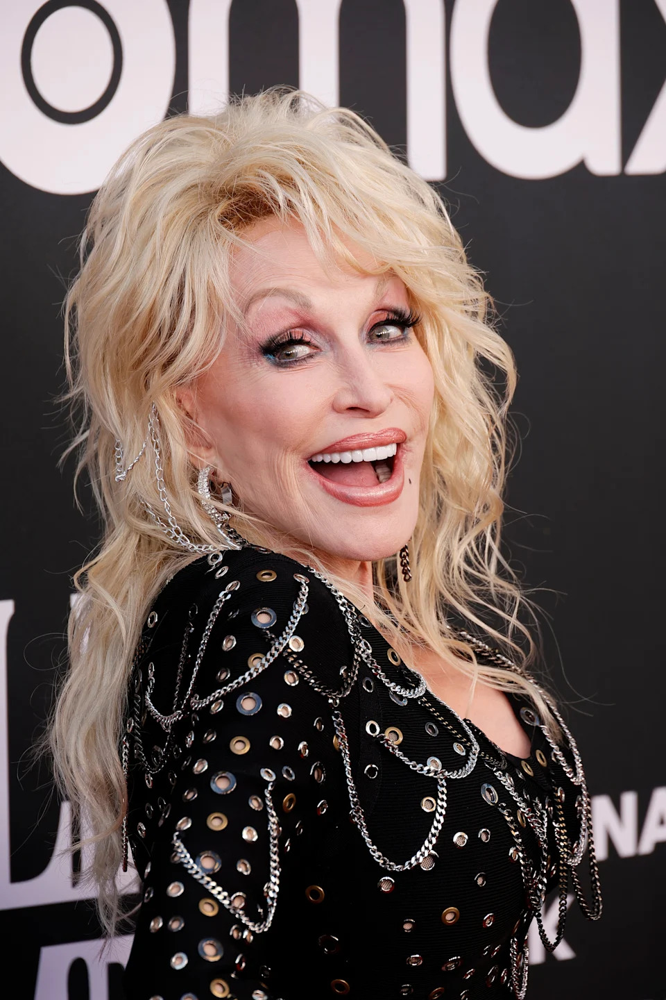 Person with long blond hair wearing an embellished black outfit, smiling at a red carpet event