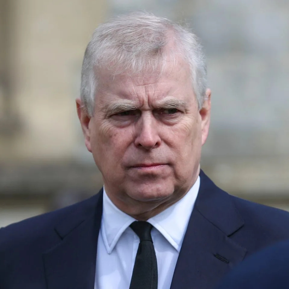  Prince Andrew wears a black suit with a white shirt and black tie and has white gray hair and is scowling. 