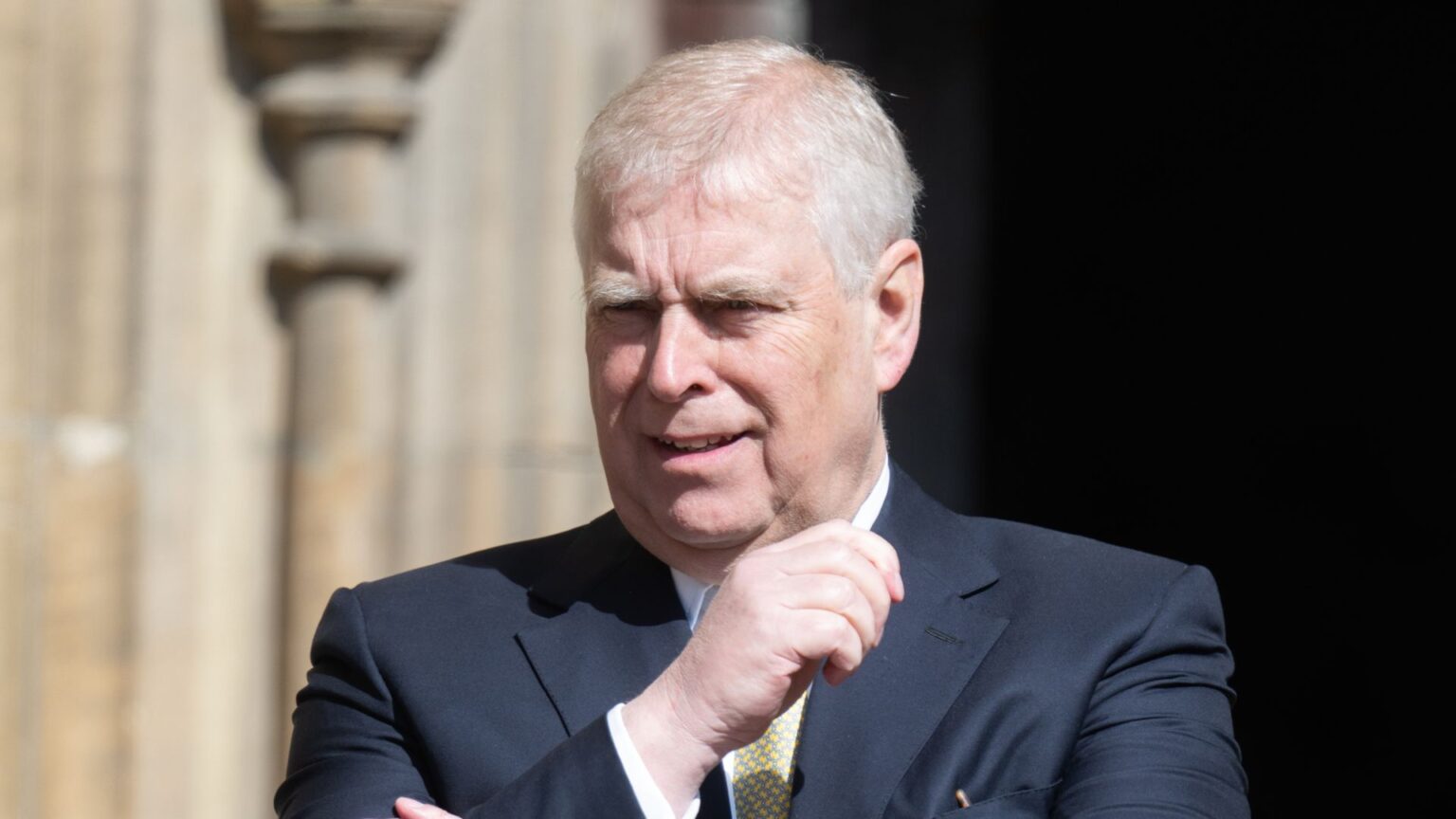 Prince Andrew looks bemused while looking at something wile wearing a suit with a white shirt and a golden tie, he has white gray hair