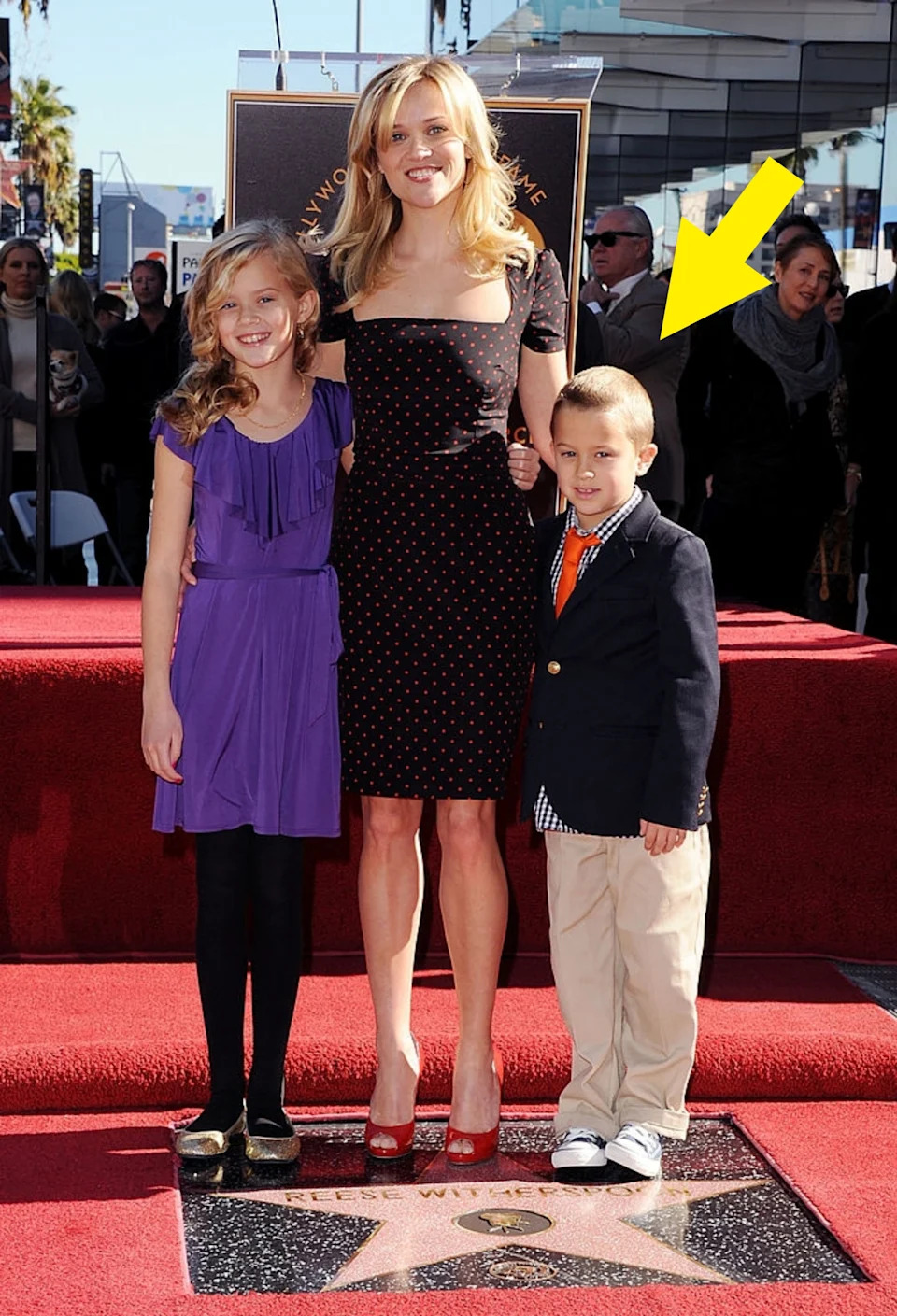 Person in a polka dot dress stands with two children on a Hollywood Walk of Fame star, surrounded by onlookers