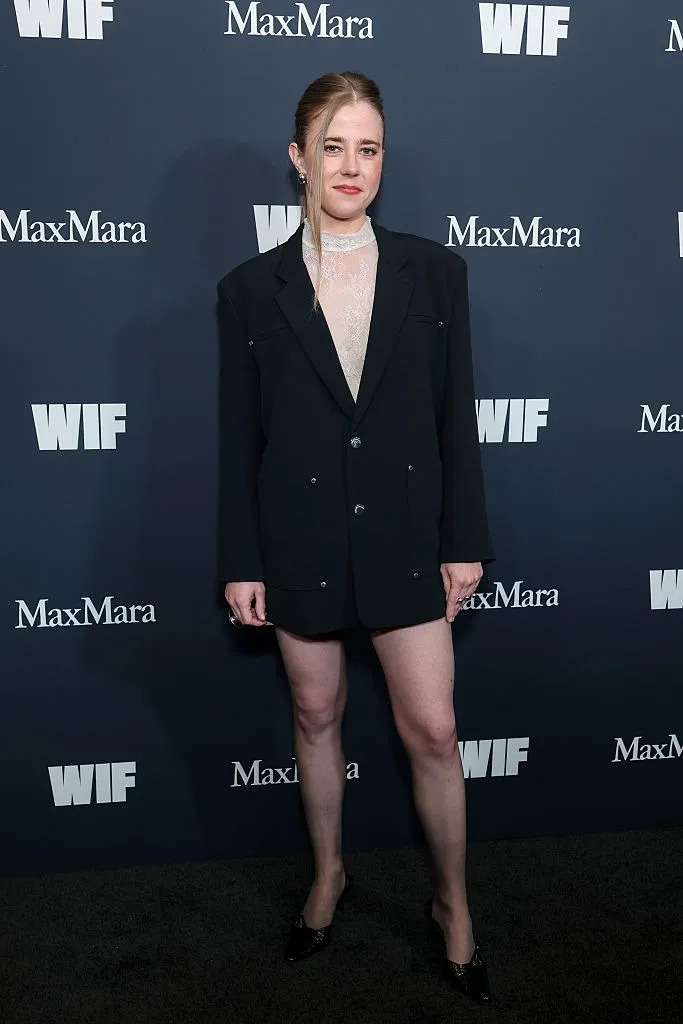 Person poses in oversized black blazer and heeled shoes at an event backdrop featuring "MaxMara" and "WIF" logos