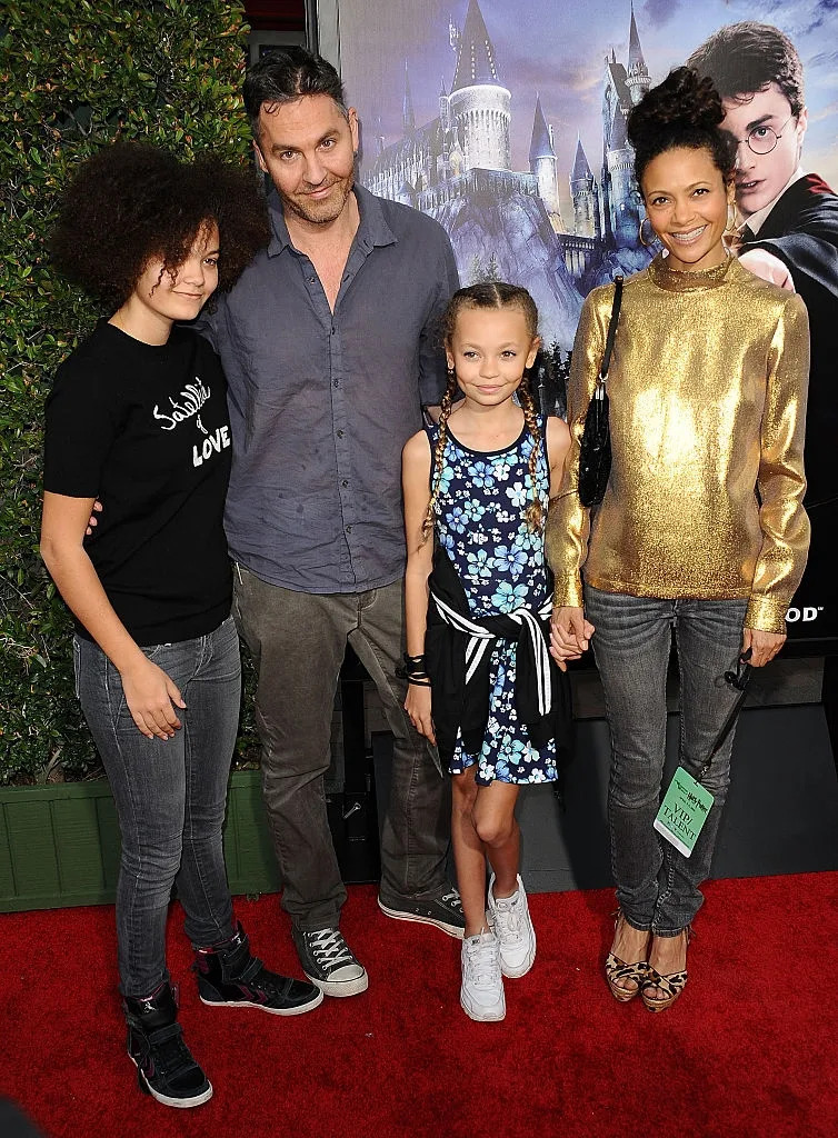 Group of four people posing on a red carpet; a woman in a gold top, a man, a teen in casual clothes, and a child in a floral dress