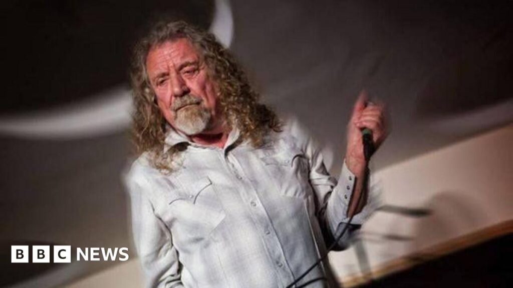 Music venue says surprise Robert Plant gig was ‘amazing’ A man with long curly dark blond hair and a grey beard wearing a white and grey shirt, holds a microphone in his left hand.