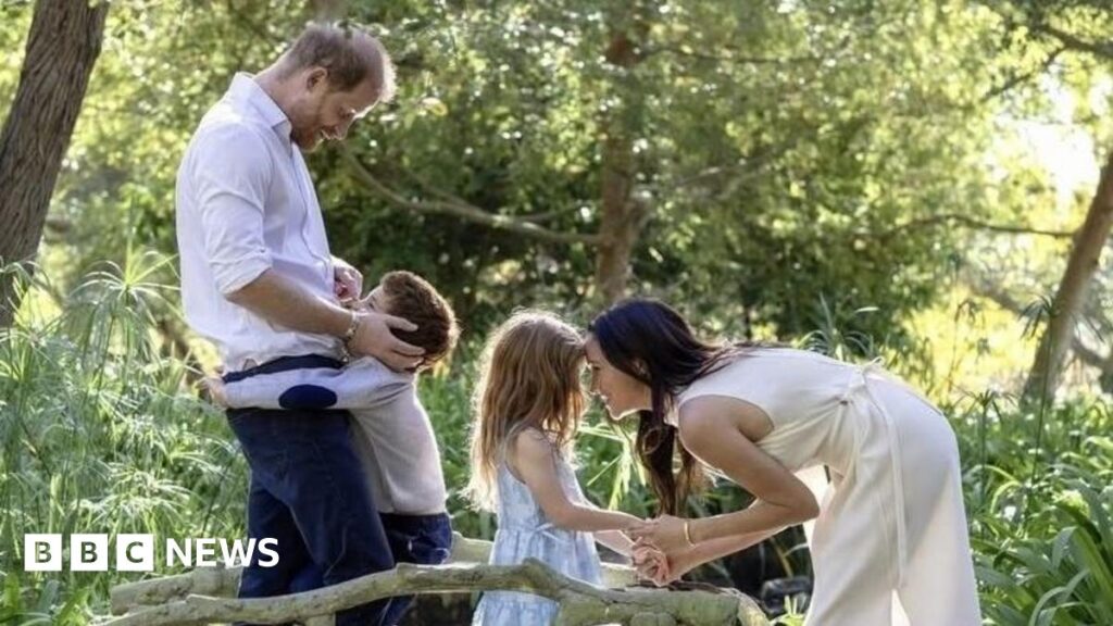 Archie is hugging his father, Prince Harry, the Duke of Sussex, while Meghan is bent to eye-level and holding hands with Lilibet, as they all stand on what appears to be carved mini wooden bridge over a brook among trees.