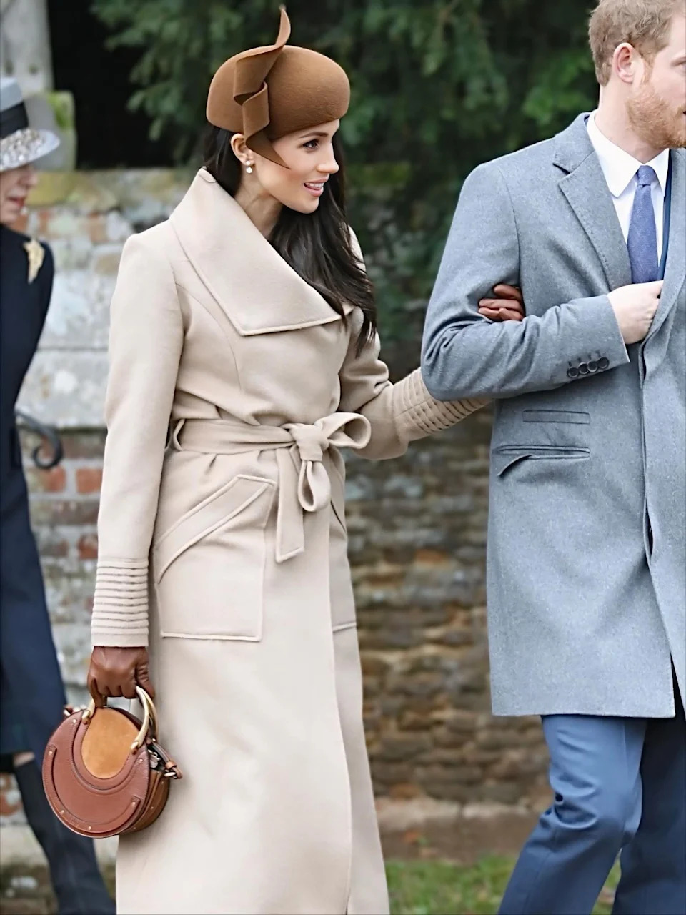Meghan Markle wearing a beige Sentaler wrap coat and brown fascinator while walking with Prince Harry.