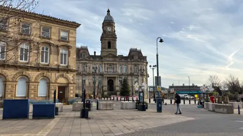 Gemma Dillon/BBC Image shows Dewsbury town centre. 
The town hall is in the background, a big stone building.
To the left of shot is also a boarded up , stone building which used to be Barclays bank but has stood empty for the last two years. 
