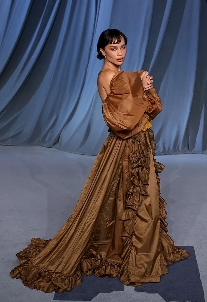 Person in elegant off-the-shoulder gown with ruffled details stands against a draped backdrop