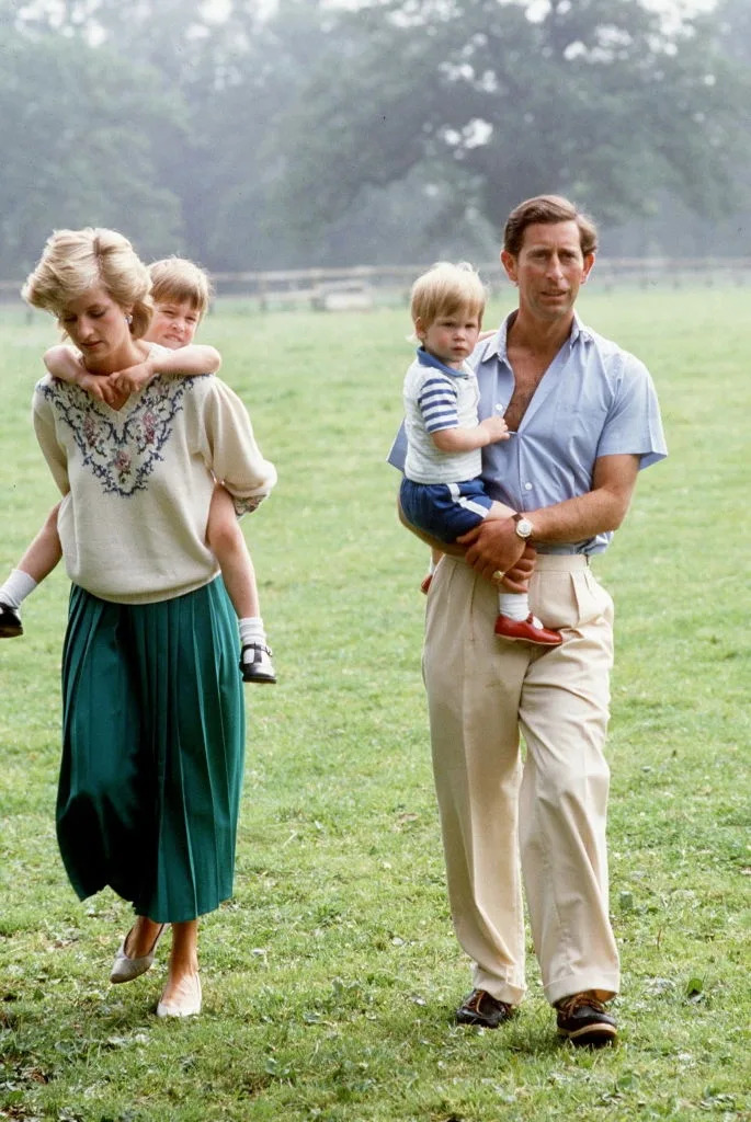 A woman in a long skirt carries a child on her back, while a man in a short-sleeved shirt holds another child. They're walking on grass outdoors