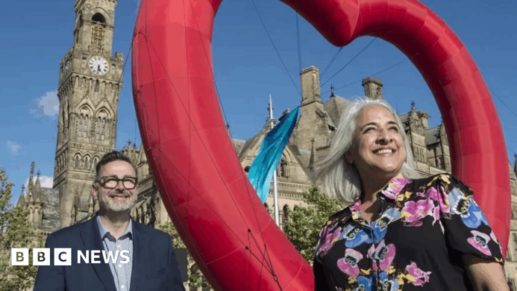 New Year Honours for Bradford City of Culture pioneers Dan Bates, wearing a blue jacket, stands to the left of the image while Shanaz Gular, wearing a patterned shirt, stands on the right. A red heart is between them with Bradford City Hall behind them.