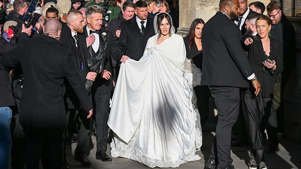 Holly Ramsay entering the church surrounded by security staff. She is wearing a white cape over her white wedding dress.