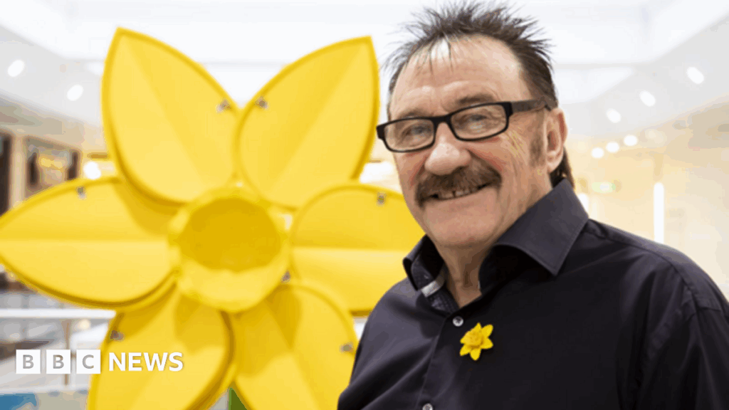 Chuckle Brother and Roy Clarke hailed in New Year Honours A man has black, short spiky hair, a moustache and black framed glasses. He is wearing a black shirt with a yellow daffodil pin. In the background is a large daffodil structure