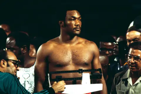 Getty Images George Foreman is standing, shirtless, on a weighing scale during a weigh-in event. He is surrounded by several people, some partially visible, including a man operating the scale. The background is dark, with the crowd and event setting creating a focused atmosphere typical of a pre-fight weigh-in.