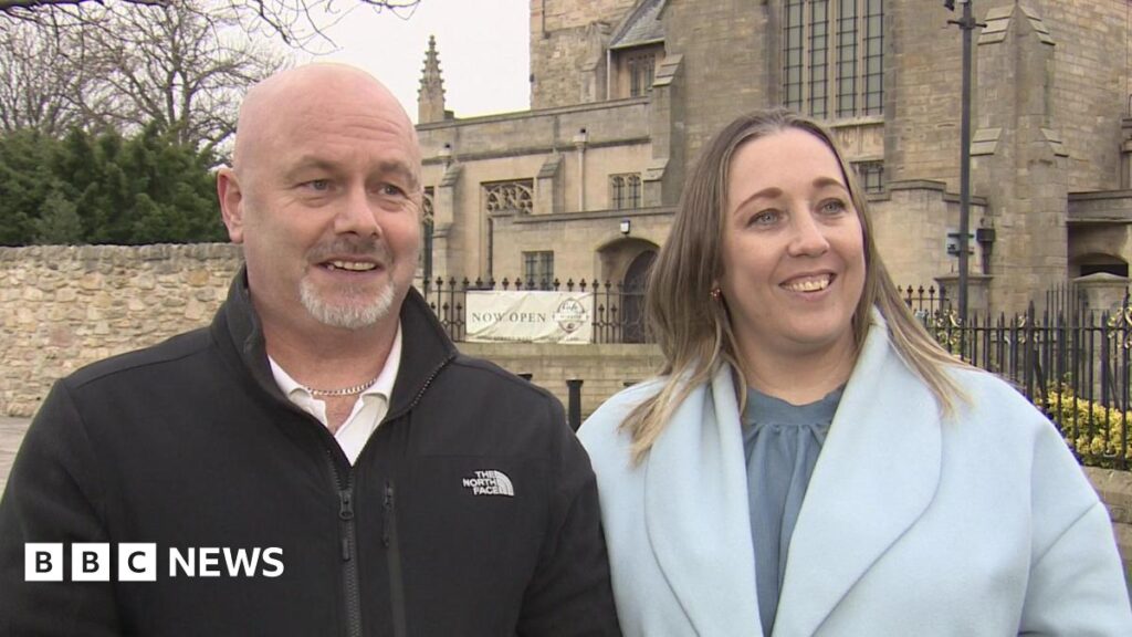 New Year’s Honours for North East knife crime campaigners Simon and Tanya Brown standing side by side outside a large sandstone building with decorated windows and spires. Mr Brown is bold and has a grey goatee. He is wearing a black zip-up fleece over a whites shirt and he is wearing a silver chain around his neck. Mrs Brown has shoulder-length light brown hair and is wearing a baby blue coat over a grey-blue blouse. They are speaking to a reporter who is interviewing them out of shot.