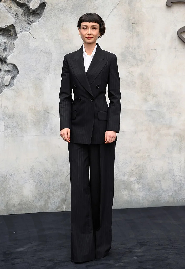 Person in a tailored dark suit and white shirt, standing against a textured backdrop
