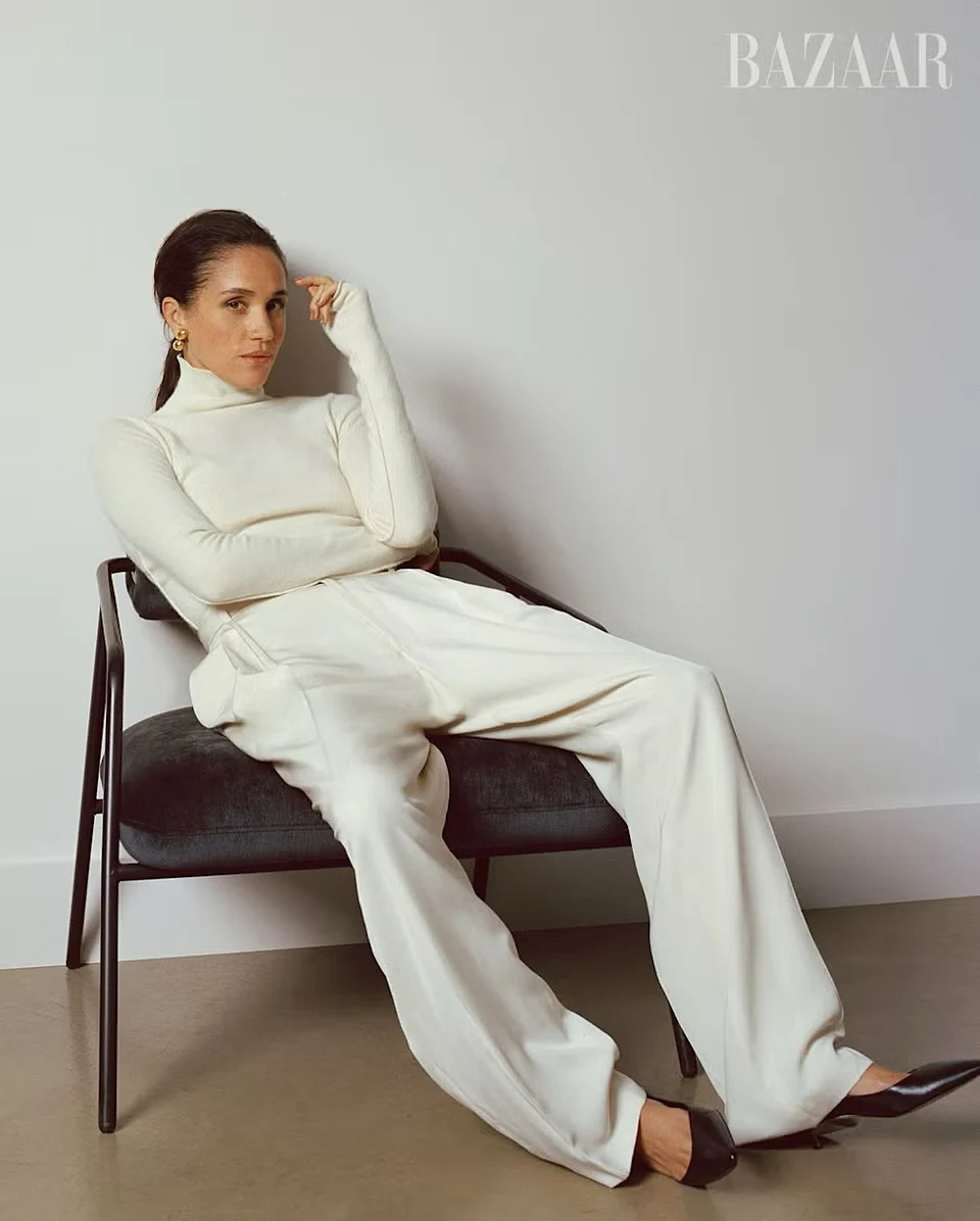 Meghan Markle seated on a chair wearing a white turtleneck and matching trousers in a minimalist studio setting.