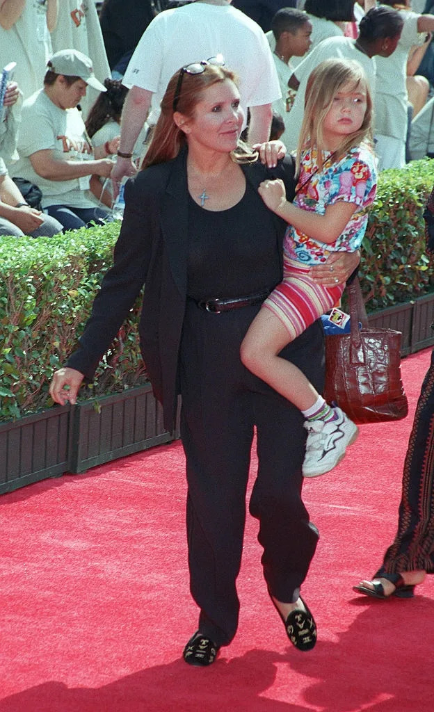 Person in black suit holding child wearing colorful outfit on a red carpet