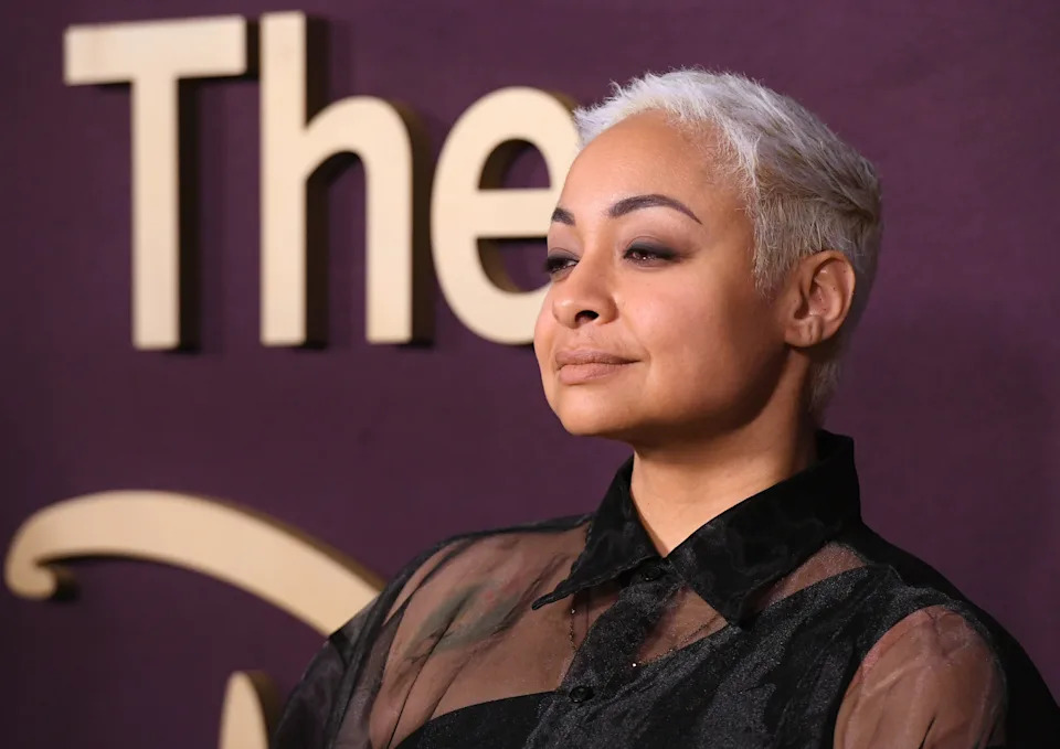 Raven-Symone with short hair in a sheer top poses confidently on a red carpet with a logo backdrop