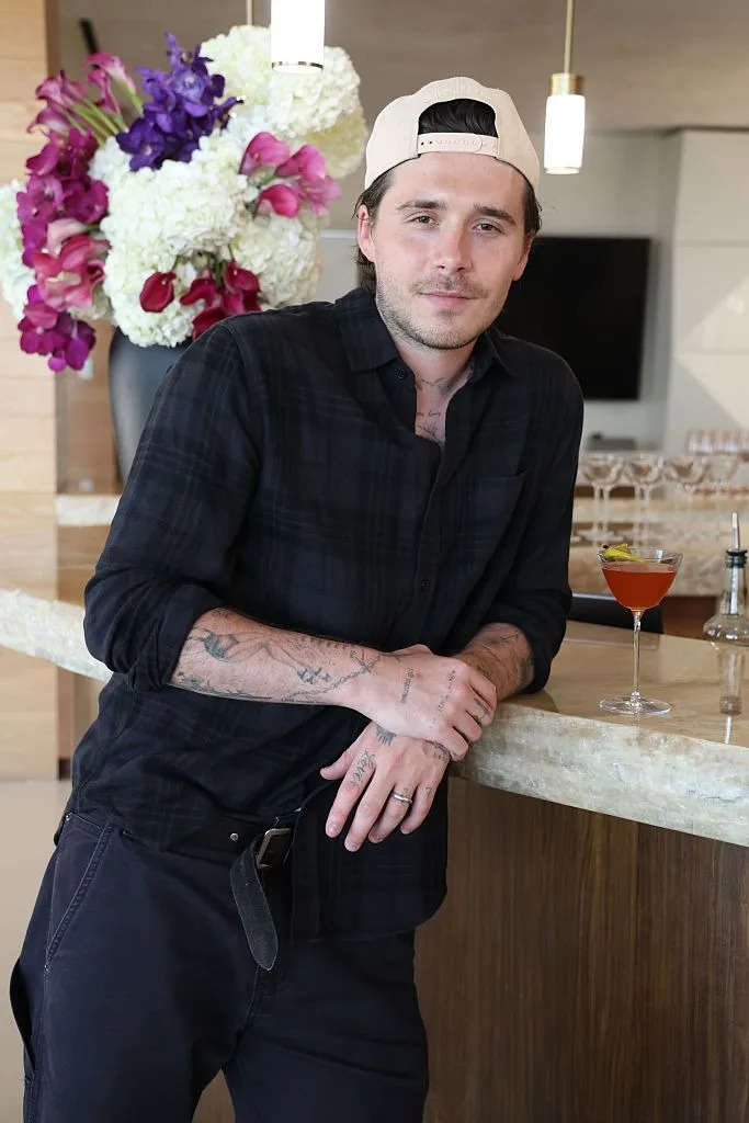 Person in casual shirt and cap leans on a bar, with cocktail and vibrant floral arrangement in background