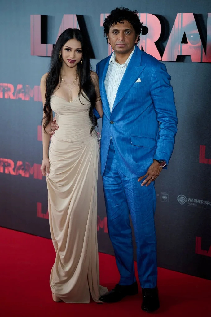 Two individuals pose on a red carpet. The man wears a blue suit, and the woman wears a form-fitting cream dress
