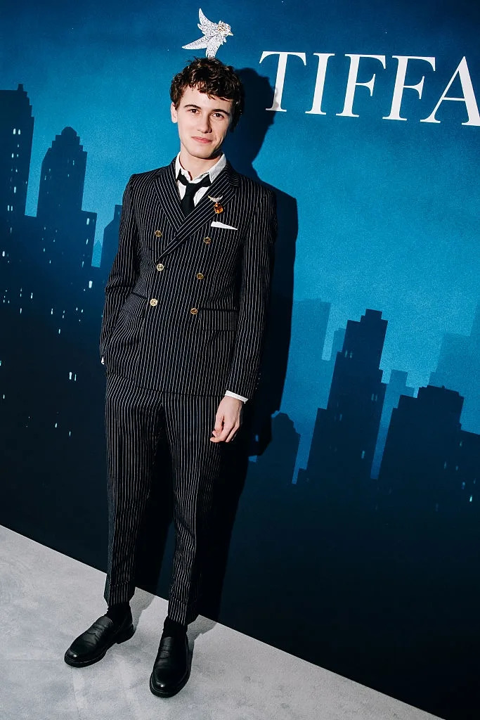 A person in a pinstripe suit stands at an event, posing in front of a backdrop with city skyline silhouettes