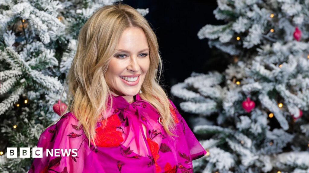Kylie Minogue poses in a pink shirt next to a pair of Christmas Trees