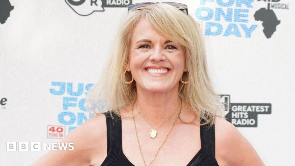 Coronation Street star dedicates MBE to working class actors Sally Lindsay smiles at the camera. She has long blonde hair and wears gold hoop earrings two gold necklaces and a black sleeveless dress.