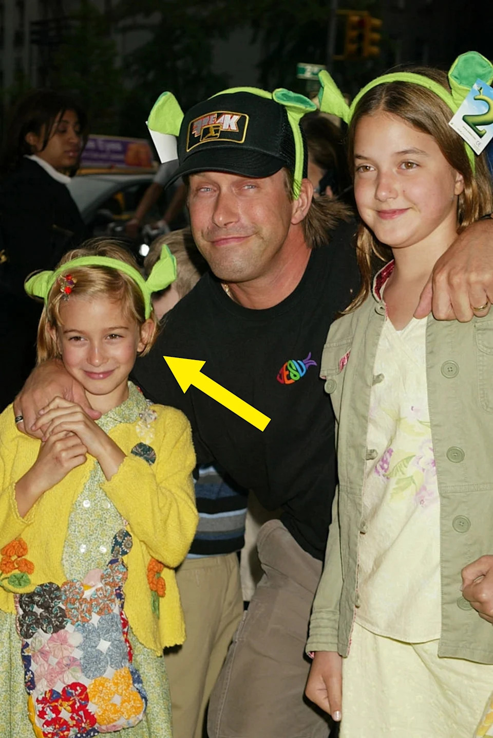 A person with two children wearing Shrek headbands on a city street, smiling for a photo