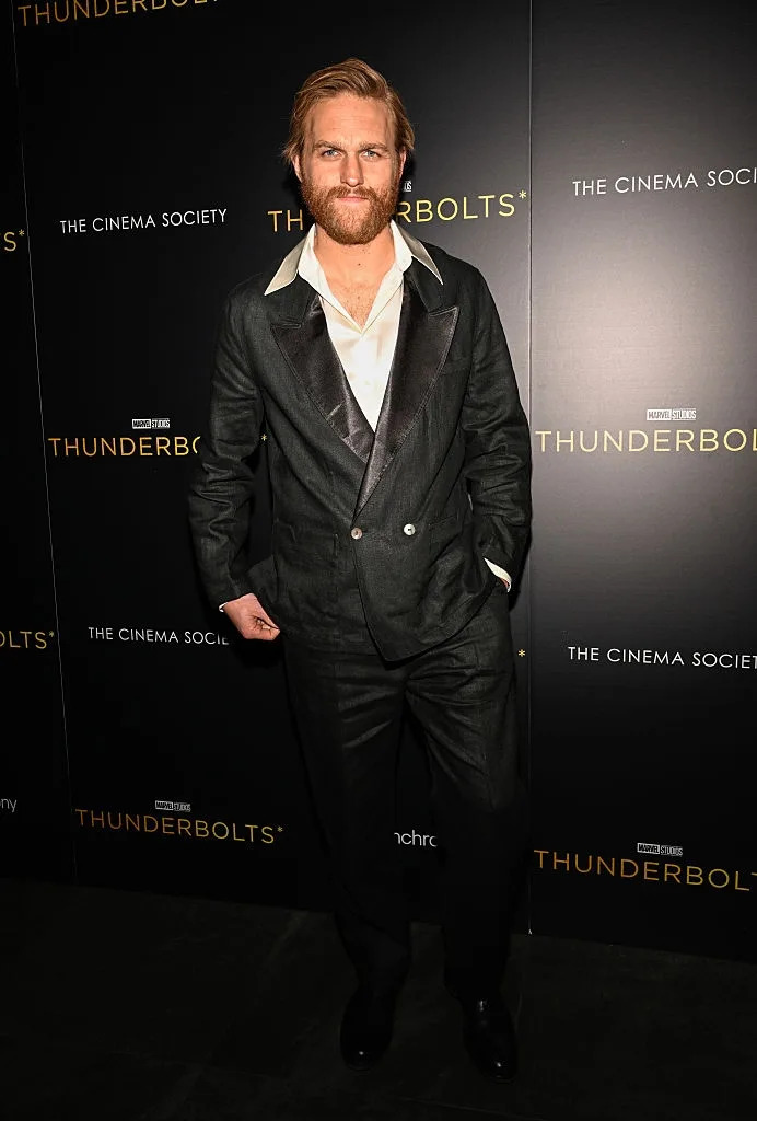 Person on the red carpet in a tailored suit with a white shirt, posing for photos at a "Thunderbolts" event backdrop