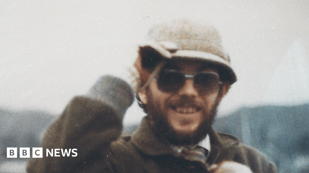 Kenner Elias Jones is a white man in his 30s or 40s in this picture, with a brown beard and moustache. He wears dark lensed prescription glasses and a cream coloured hat which he is holding, a checked shirt and tie covered by a brown coat with a collar. He is smiling at the camera. The image is grainy.