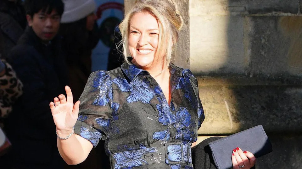 A blonde woman smiles outside a church. She is dressed for a wedding at which she is in a black and blue dress.